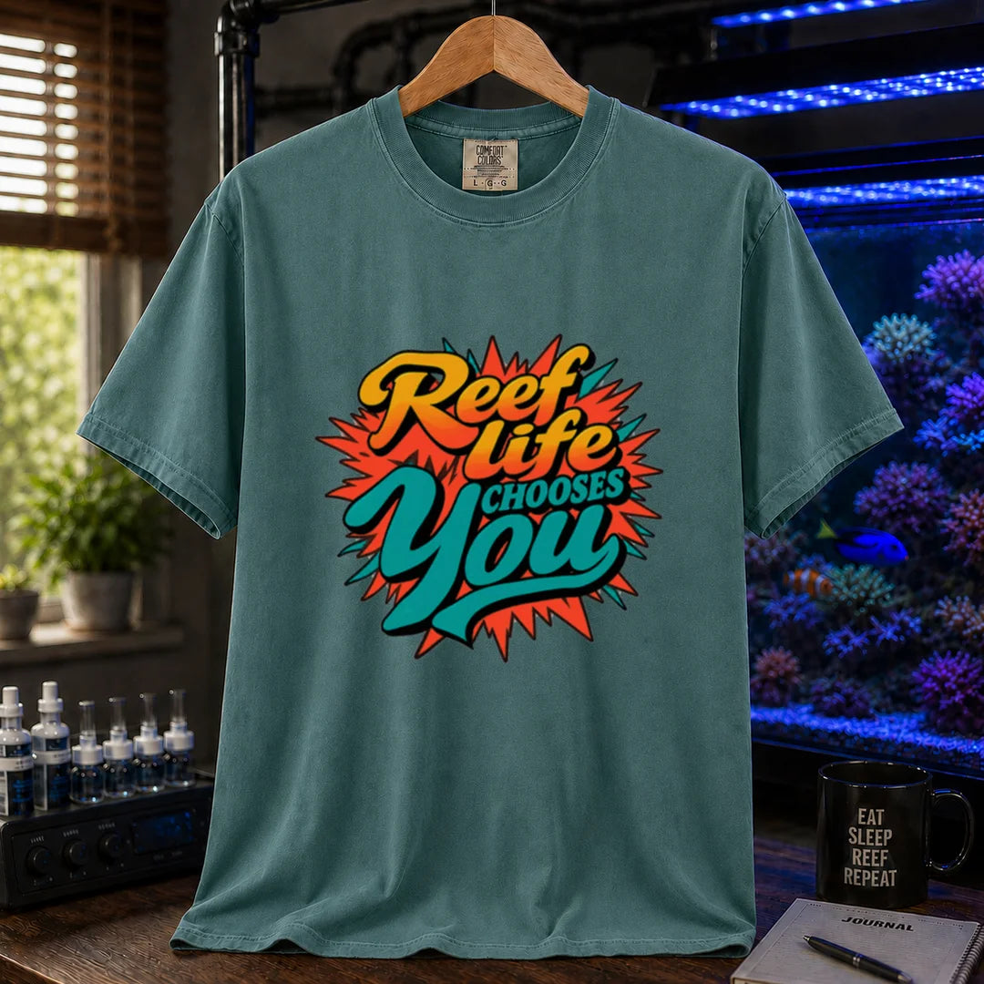Saltwater aquarium graphic t-shirt featuring vibrant yellow, orange, and teal retro pop-art typography reading Reef Life Chooses You with a red starburst design printed on a green tee displayed in a reef room setting