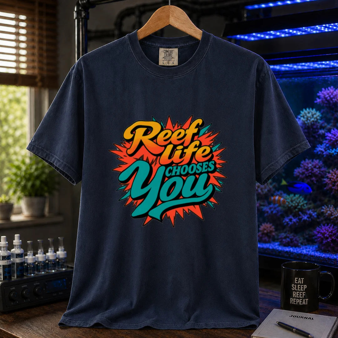 Saltwater aquarium graphic t-shirt featuring vibrant yellow, orange, and teal retro pop-art typography reading Reef Life Chooses You with a red starburst design printed on a navy tee displayed in a reef room setting