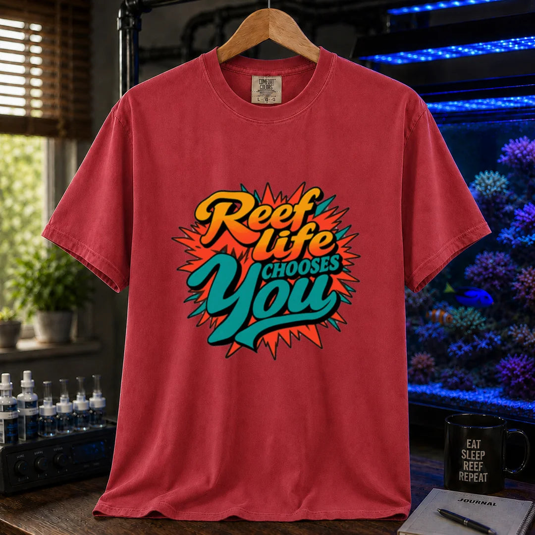 Saltwater aquarium graphic t-shirt featuring vibrant yellow, orange, and teal retro pop-art typography reading Reef Life Chooses You with a red starburst design printed on a red tee displayed in a reef room setting