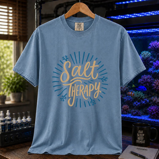 Saltwater aquarium graphic t-shirt featuring textured cream script typography reading Salt Therapy with blue sunburst rays and coral accents printed on a blue tee displayed in a reef room setting