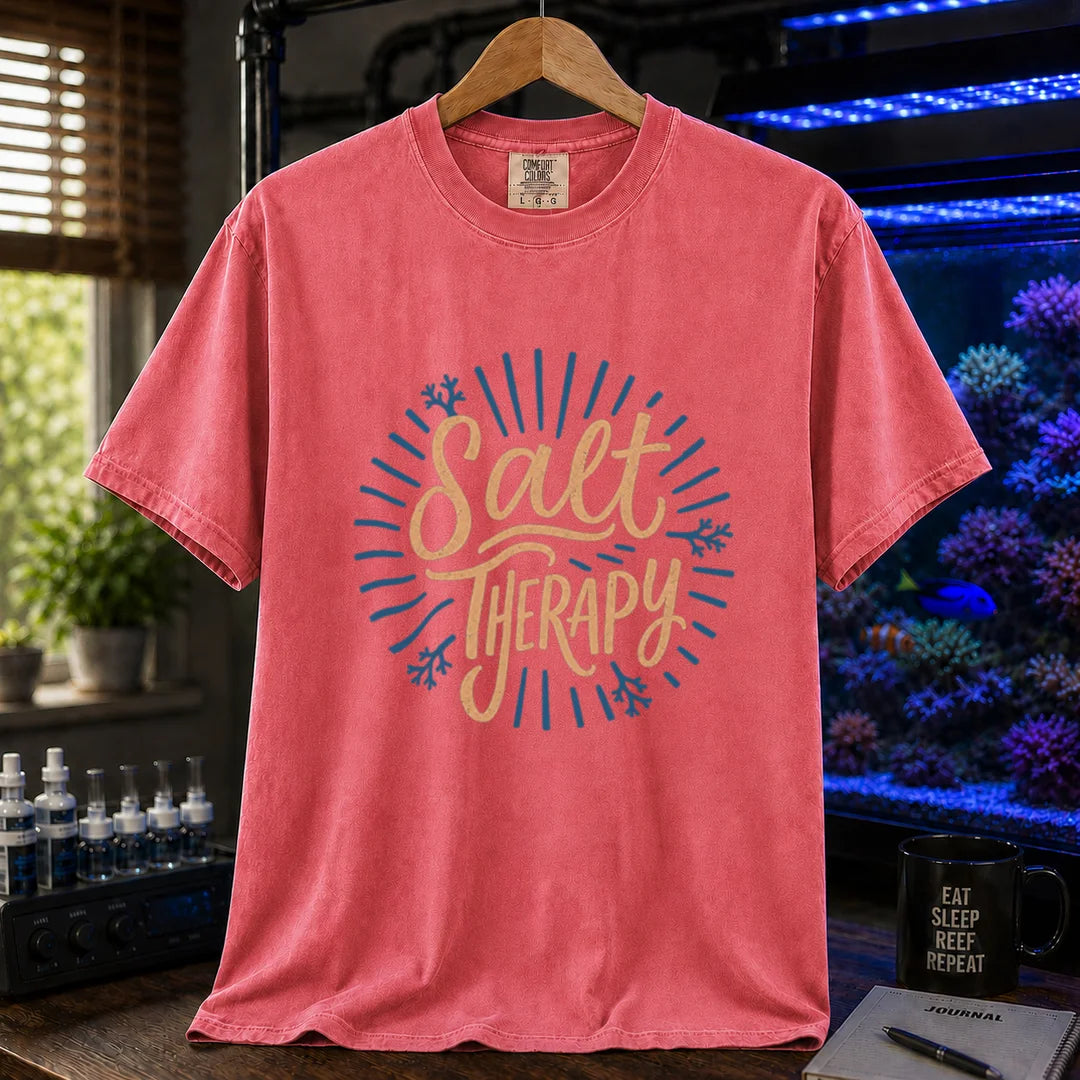 Saltwater aquarium graphic t-shirt featuring textured cream script typography reading Salt Therapy with blue sunburst rays and coral accents printed on a watermelon tee displayed in a reef room setting