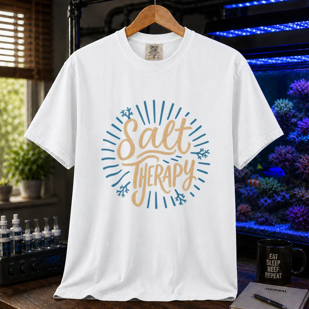 Saltwater aquarium graphic t-shirt featuring textured cream script typography reading Salt Therapy with blue sunburst rays and coral accents printed on a white tee displayed in a reef room setting
