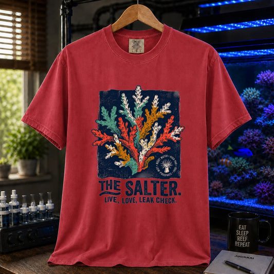Saltwater aquarium graphic t-shirt featuring a multi-color coral illustration and bold navy typography reading The Salter. Live, Love, Leak Check. printed on a red tee displayed in a reef room setting