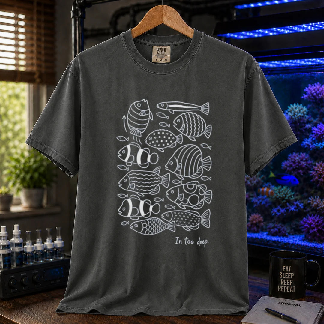 Saltwater aquarium graphic t-shirt featuring a minimalist white line-art pattern of various reef fish and the caption In too deep. printed on a dark grey tee displayed in a reef room setting
