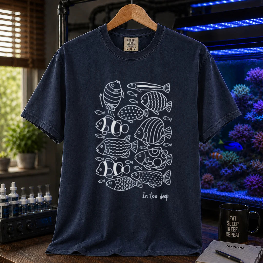 Saltwater aquarium graphic t-shirt featuring a minimalist white line-art pattern of various reef fish and the caption In too deep. printed on a navy tee displayed in a reef room setting