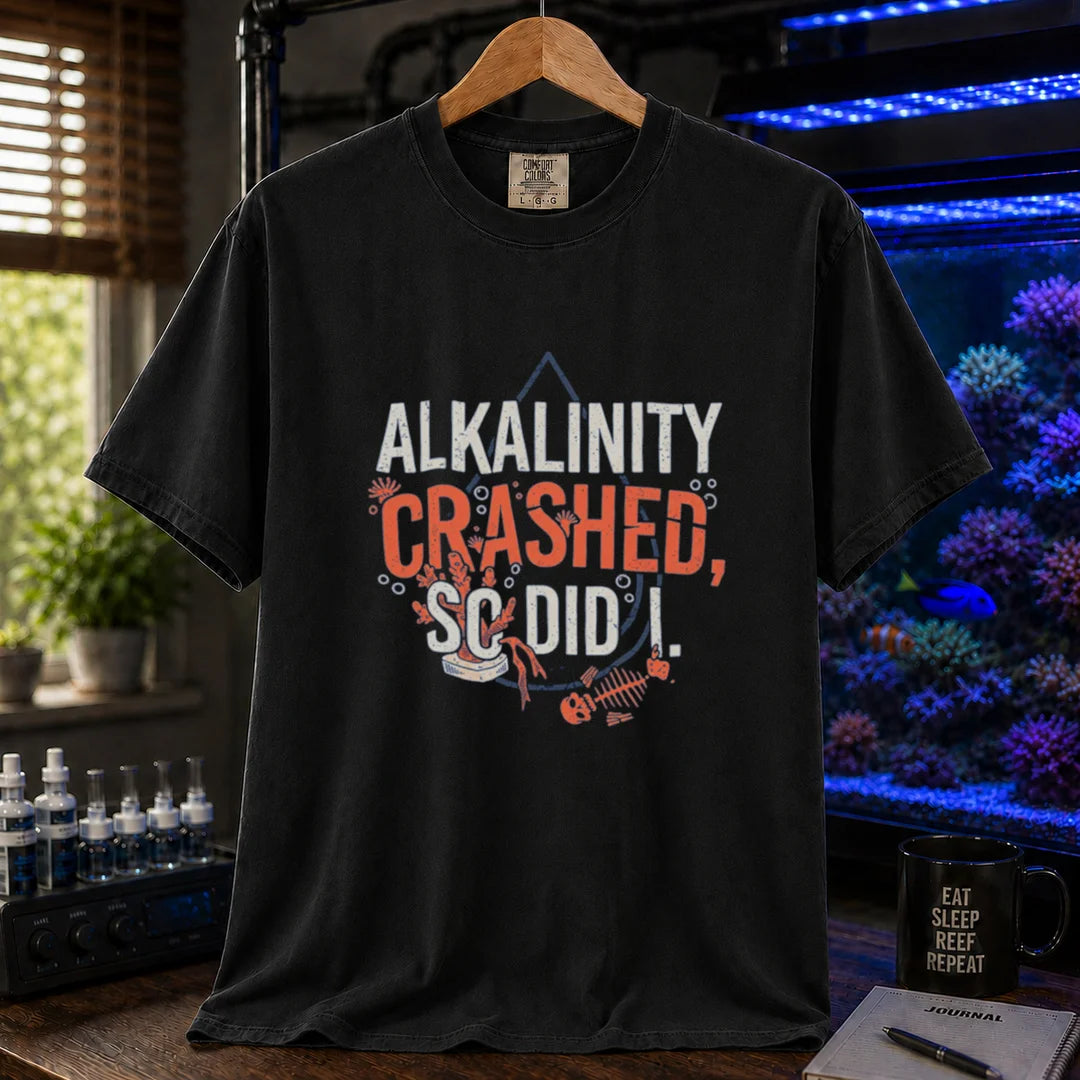 Saltwater aquarium graphic t-shirt featuring distressed white and salmon typography reading Alkalinity Crashed, So Did I. with a water drop outline, coral, and a fish skeleton printed on a black tee displayed in a reef room setting
