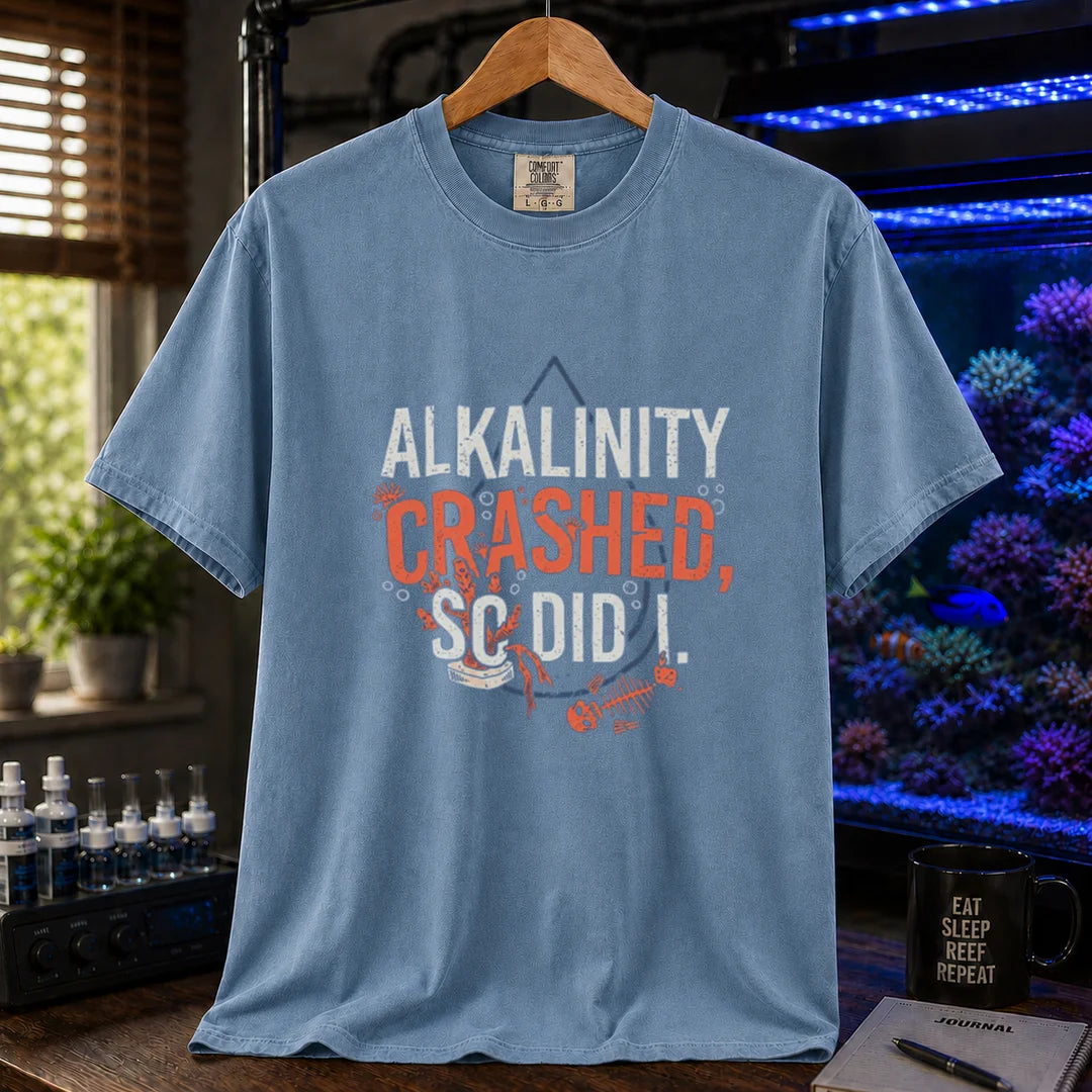 Saltwater aquarium graphic t-shirt featuring distressed white and salmon typography reading Alkalinity Crashed, So Did I. with a water drop outline, coral, and a fish skeleton printed on a blue tee displayed in a reef room setting