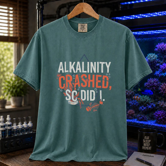 Saltwater aquarium graphic t-shirt featuring distressed white and salmon typography reading Alkalinity Crashed, So Did I. with a water drop outline, coral, and a fish skeleton printed on a green tee displayed in a reef room setting
