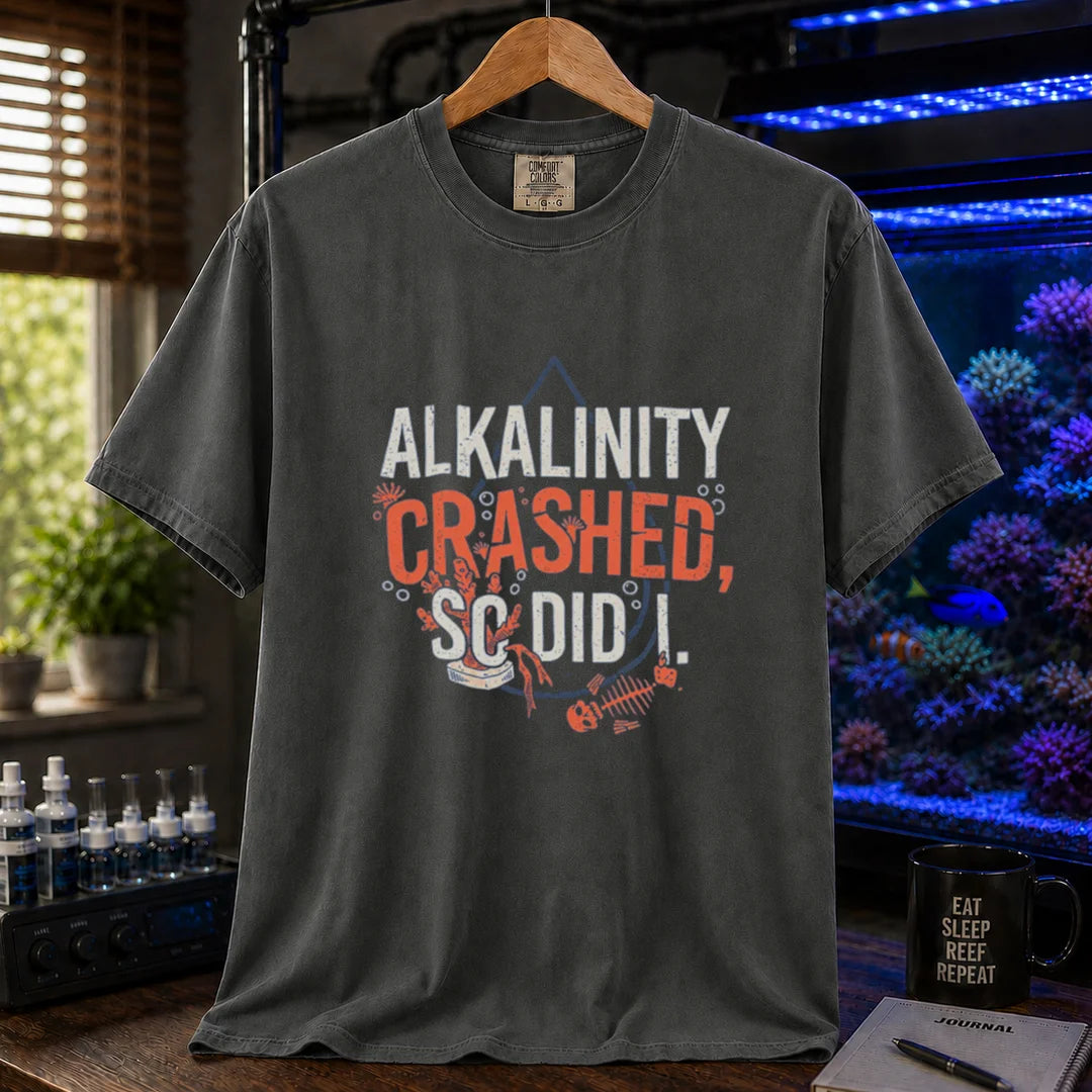 Saltwater aquarium graphic t-shirt featuring distressed white and salmon typography reading Alkalinity Crashed, So Did I. with a water drop outline, coral, and a fish skeleton printed on a dark grey tee displayed in a reef room setting