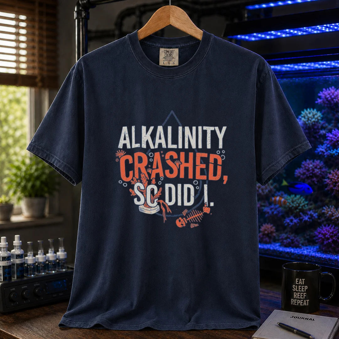 Saltwater aquarium graphic t-shirt featuring distressed white and salmon typography reading Alkalinity Crashed, So Did I. with a water drop outline, coral, and a fish skeleton printed on a navy tee displayed in a reef room setting