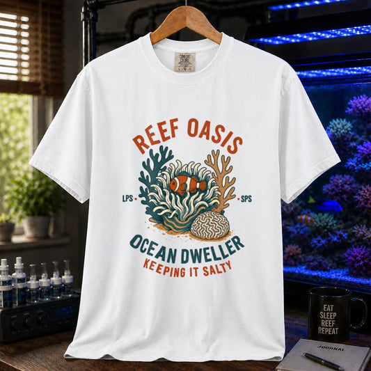 Saltwater aquarium graphic t-shirt featuring rusty-orange and teal typography reading Reef Oasis, Ocean Dweller, Keeping It Salty with an anemone, clownfish, and coral illustration printed on a white tee displayed in a reef room setting