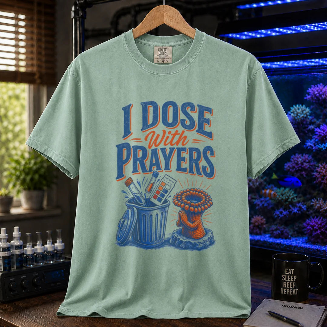 Saltwater aquarium graphic t-shirt featuring typography reading I Dose With Prayers above an illustration of test kits in a trash can and a praying coral polyp on a light green shirt