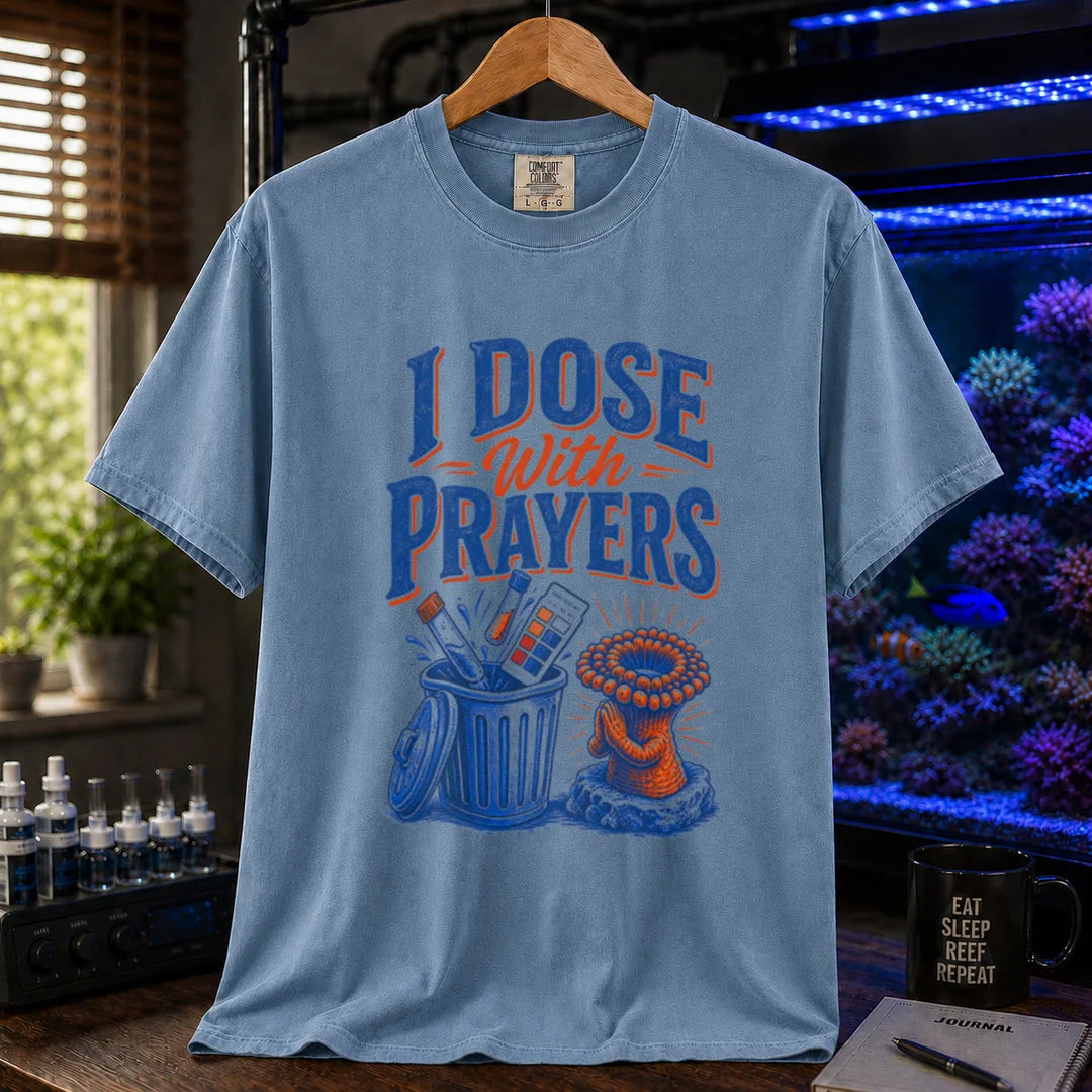Saltwater aquarium graphic t-shirt featuring typography reading I Dose With Prayers above an illustration of test kits in a trash can and a praying coral polyp on a blue shirt