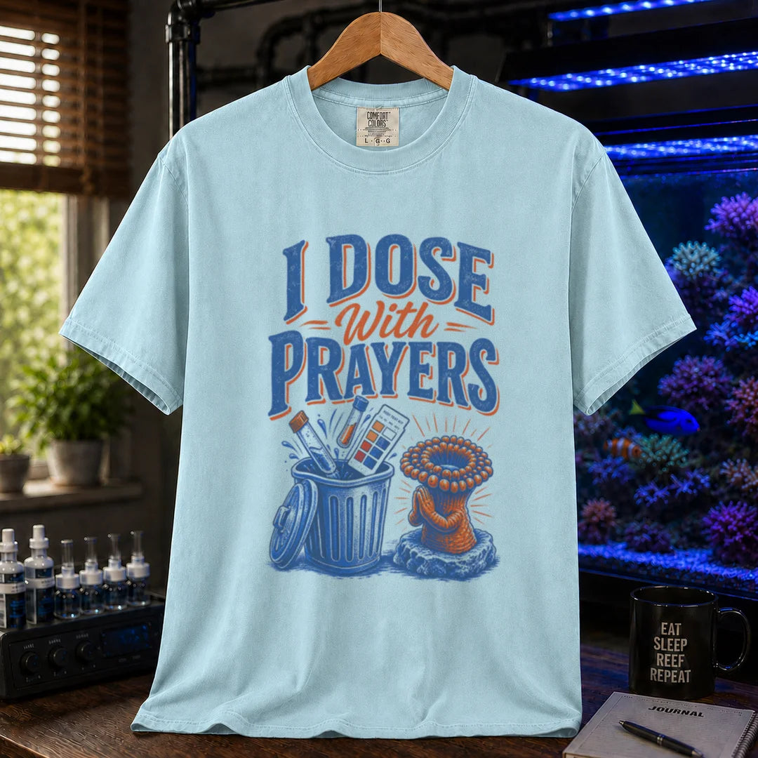 Saltwater aquarium graphic t-shirt featuring typography reading I Dose With Prayers above an illustration of test kits in a trash can and a praying coral polyp on a light blue shirt