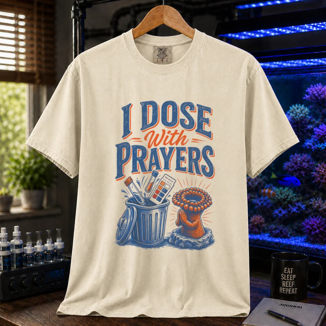 Saltwater aquarium graphic t-shirt featuring typography reading I Dose With Prayers above an illustration of test kits in a trash can and a praying coral polyp on a beige, cream, ivory shirt