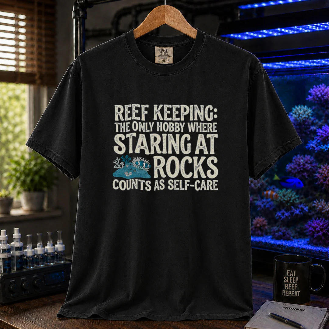 Saltwater aquarium graphic t-shirt featuring distressed typography reading Reef Keeping The Only Hobby Where Staring At Rocks Counts As Self-Care with a small rock and coral illustration on a black shirt
