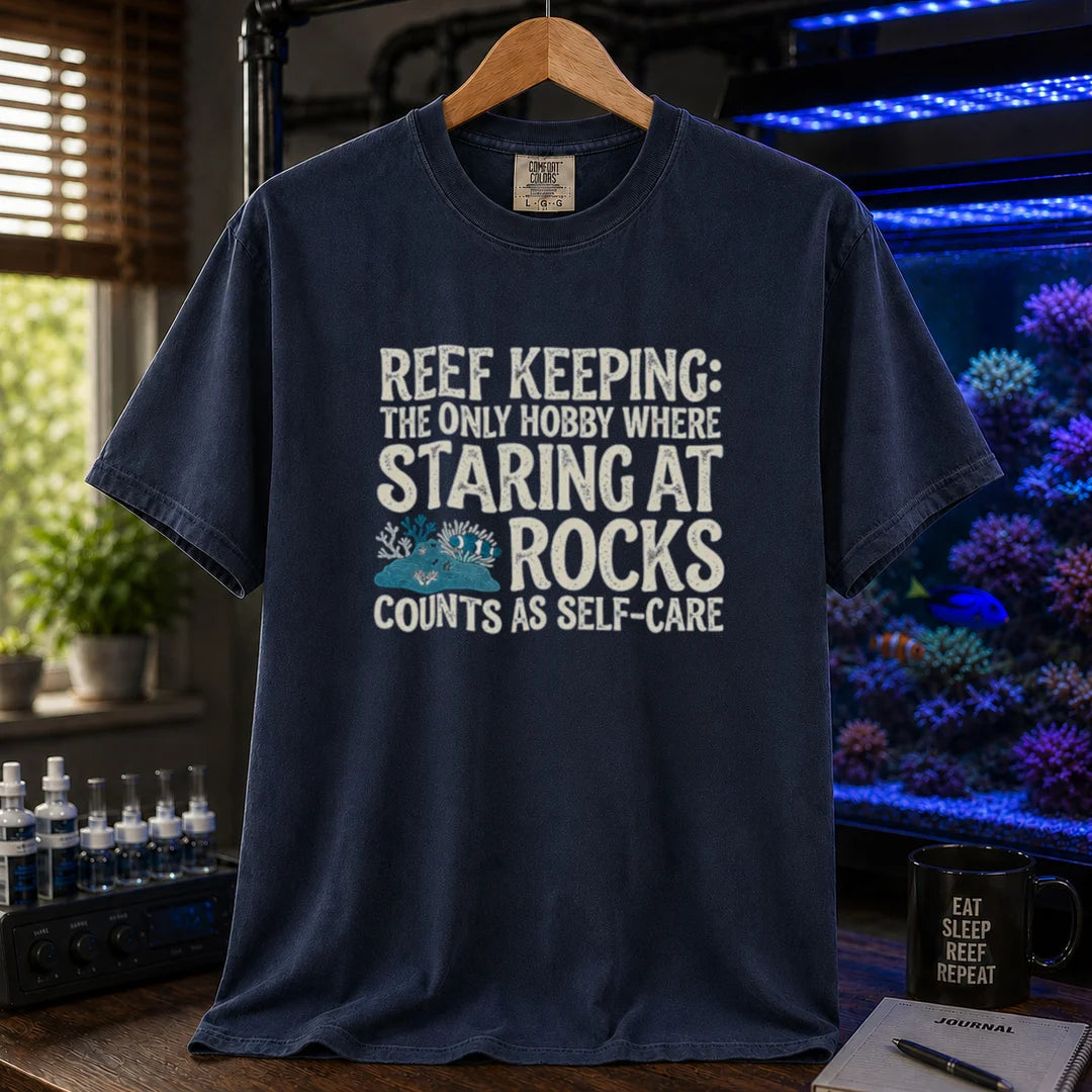 Saltwater aquarium graphic t-shirt featuring distressed typography reading Reef Keeping The Only Hobby Where Staring At Rocks Counts As Self-Care with a small rock and coral illustration on a navy shirt