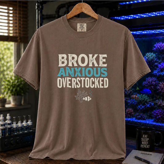 Saltwater aquarium graphic t-shirt featuring distressed typography reading Broke Anxious Overstocked with a small coral and clownfish illustration on a brown shirt