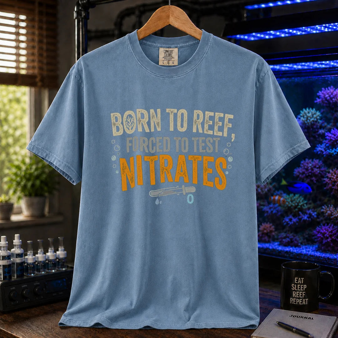 Saltwater aquarium graphic t-shirt featuring distressed cream, gray, and yellow-orange typography reading Born To Reef, Forced To Test Nitrates with a test vial icon printed on a blue tee displayed in a reef room setting